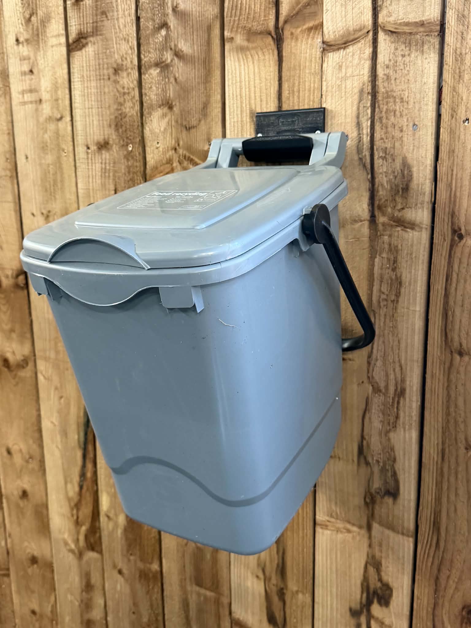 3D printed wall bracket holding a food waste caddy against a wooden fence